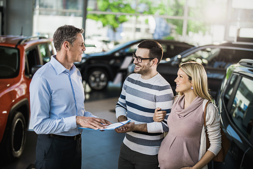 Happy pregnant couple talking to car salesperson in a showroom.
